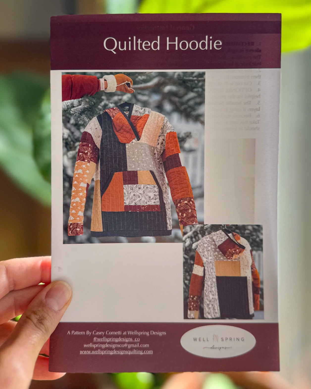 Sewing pattern with a quilted hoodie design held by a hand, with a blurred green plant in the background.