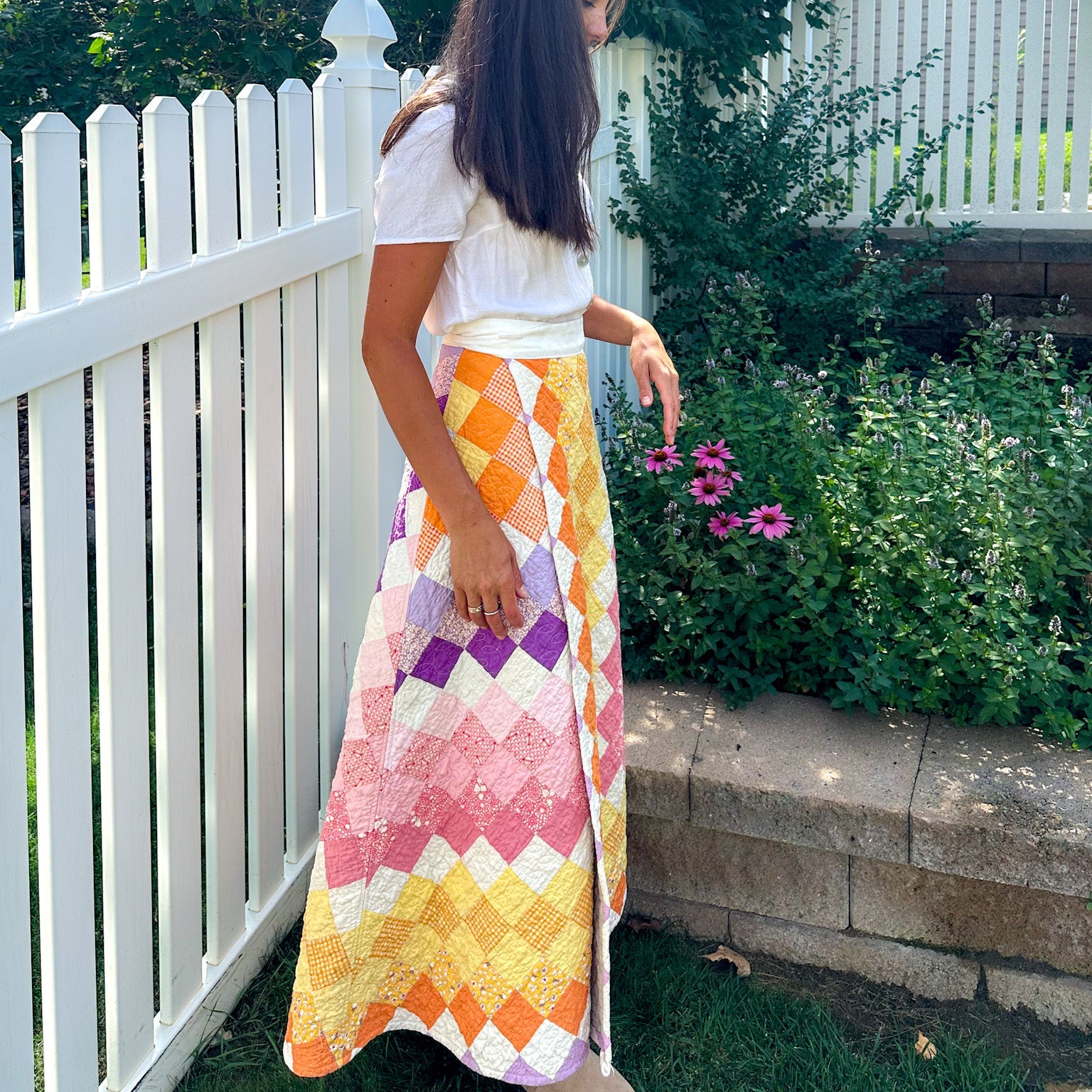 Person wearing a colorfu quilted patterned skirt standing outdoors near a white fence and greenery.
